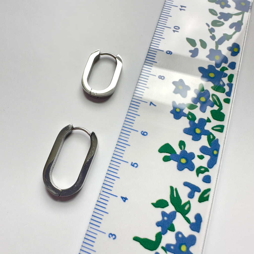 Two silver-toned oval hoop earrings displayed next to a clear plastic ruler with blue and green floral markings on a white background.