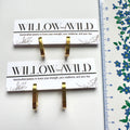 Two gold hoop earrings displayed on white branded jewelry cards, with a blue-and-white ruler along the right side for scale. The cards read WILLOW and WILD.