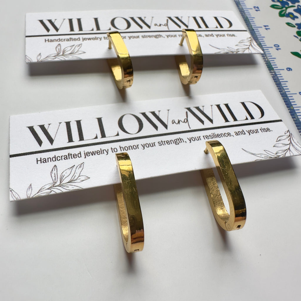 Two gold hoop earrings displayed on branded white cards, with a blue ruler visible along the top right edge on a white surface.