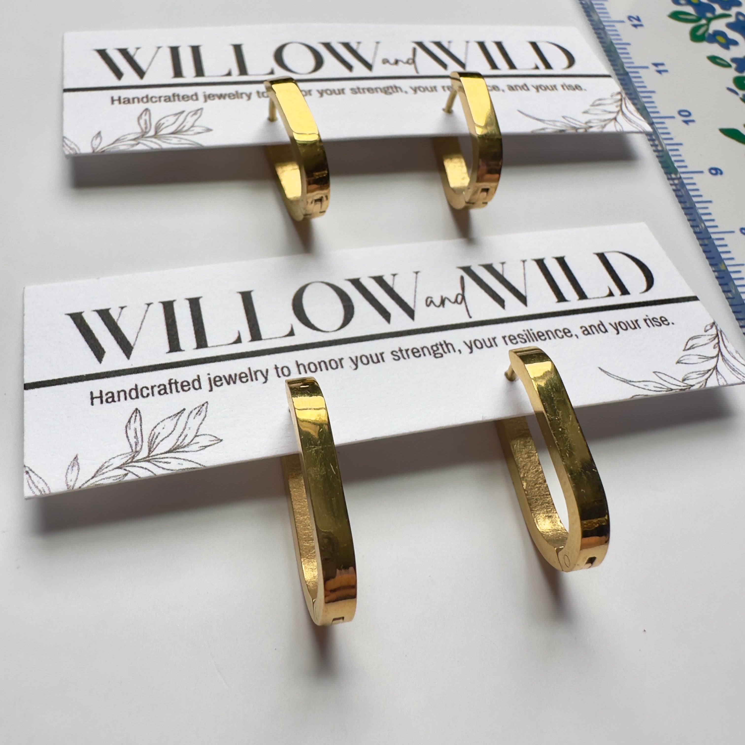 Two gold hoop earrings displayed on branded white cards, with a blue ruler visible along the top right edge on a white surface.
