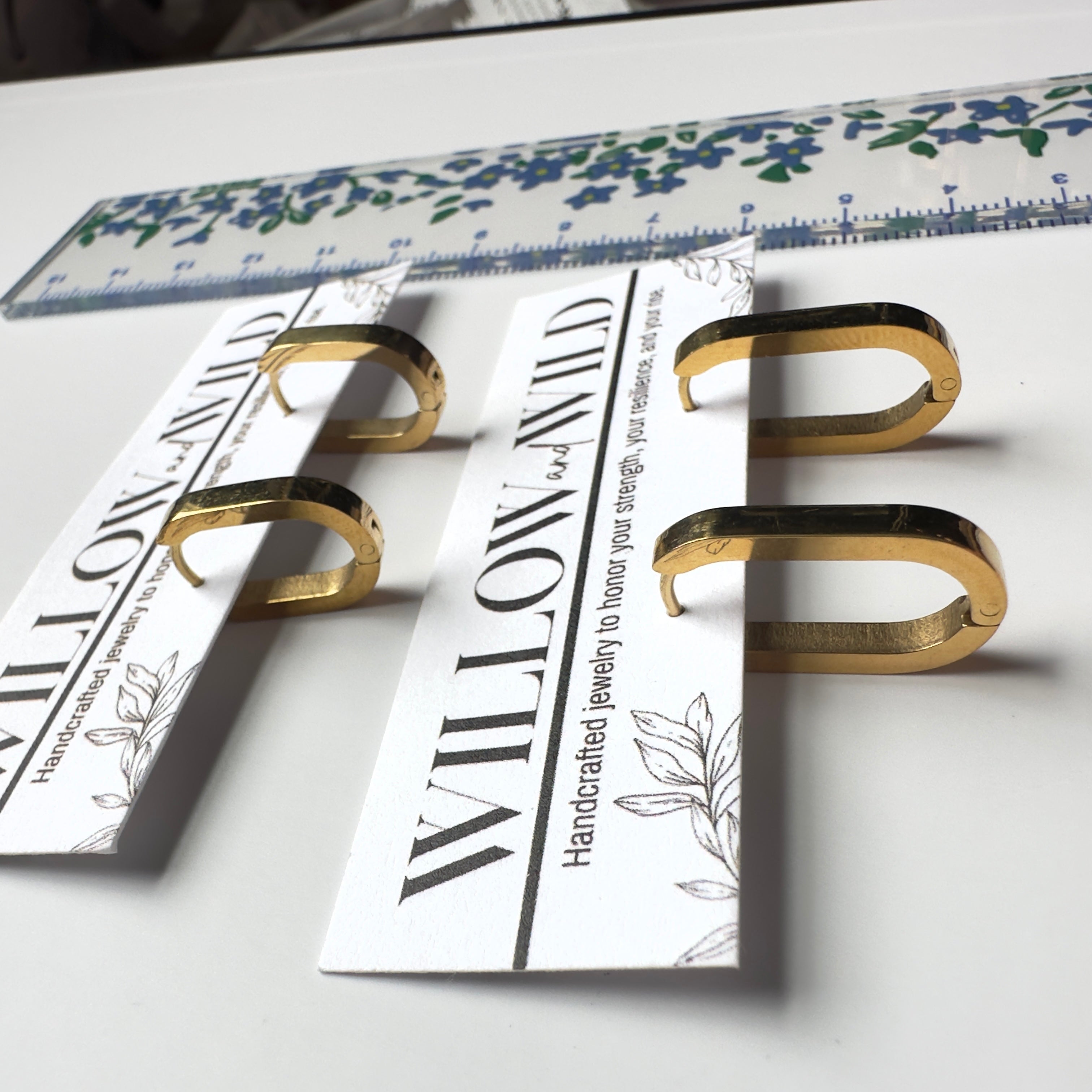 Four gold-toned metal ring stands with printed white cards, arranged on a white surface with a floral ruler in the background.